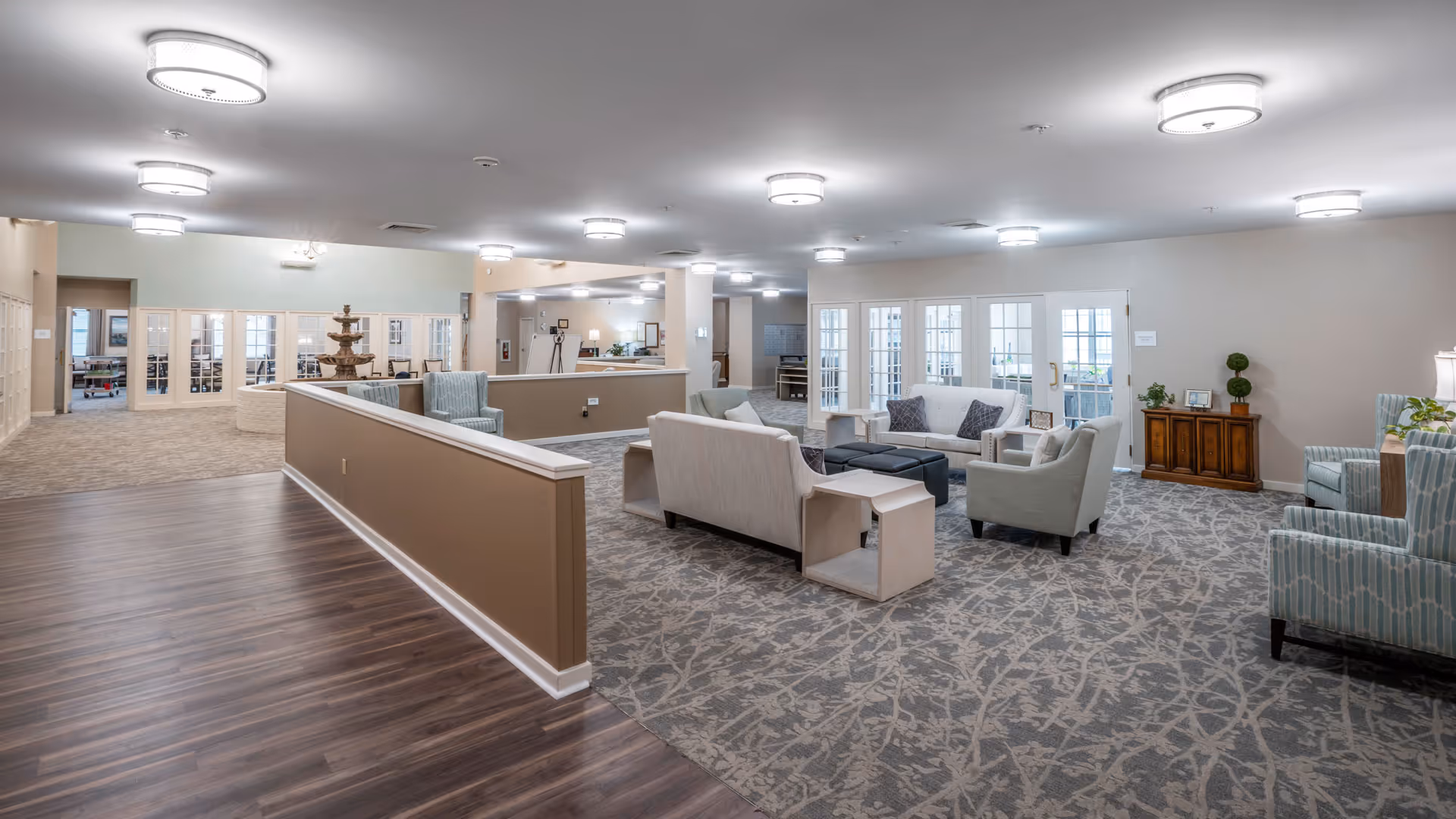 A spacious and well-lit common area in a senior living facility featuring multiple seating arrangements with sofas and armchairs on a patterned carpet. The room has light-colored walls, several ceiling lights, and a wooden floor section leading to the seating area. In the background, there are glass doors and a water fountain, with additional rooms visible through the glass panels.
