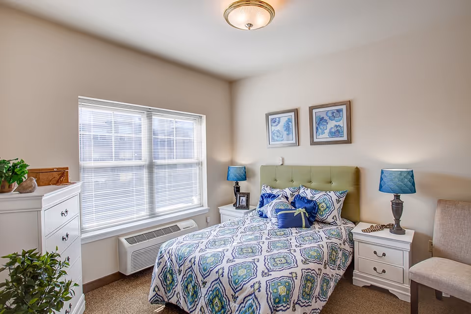 Bright furnished bedroom with a patterned bed and green tufted headboard, nightstands and lamps beside a large window.