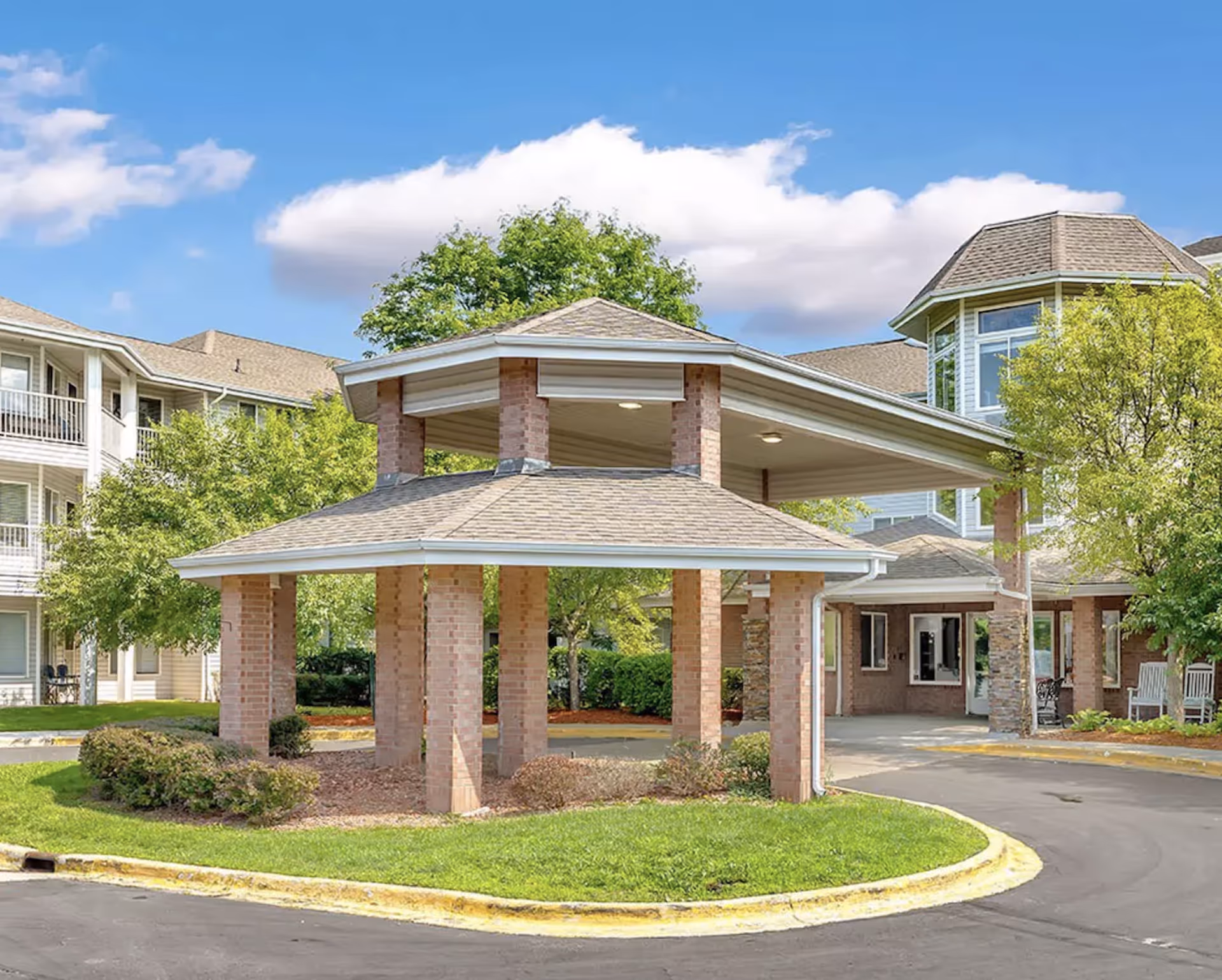 Exterior view of Independence Village of Waterford showing a covered entrance with brick pillars and a circular driveway. The building has multiple stories with balconies and large windows, surrounded by green trees and bushes under a blue sky with some clouds.