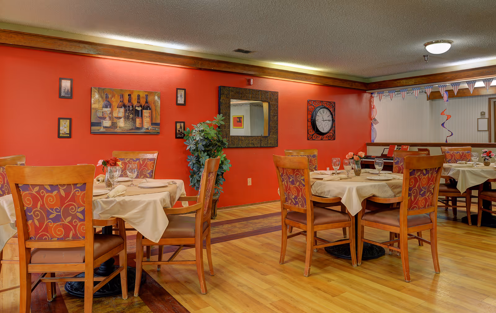 Dining room with round tables set with tablecloths and glassware, wooden chairs, and a red accent wall decorated with artwork and a mirror.
