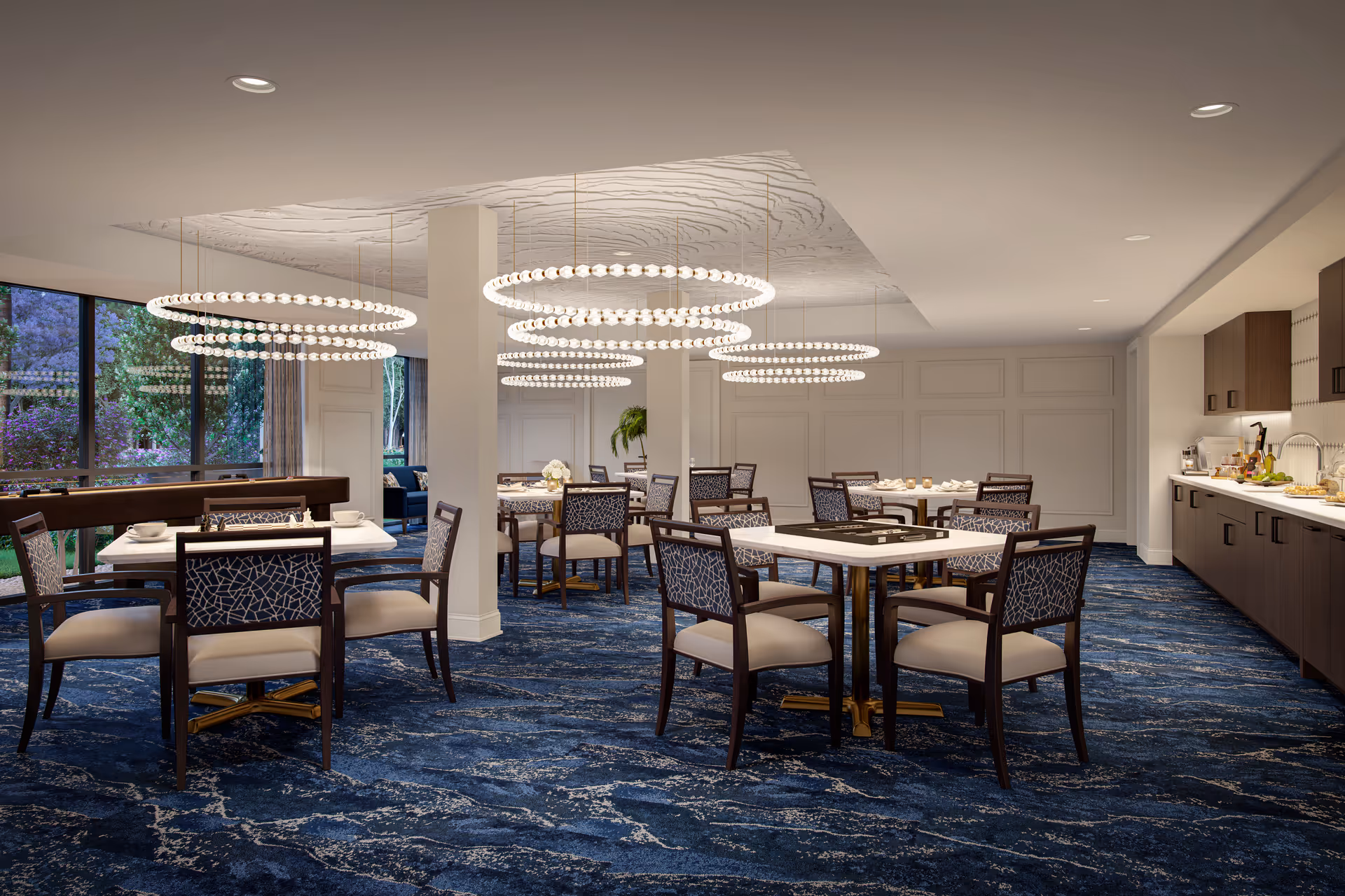 Well-lit elegant communal dining room with multiple tables and chairs, circular chandeliers, and a serving counter along one wall.