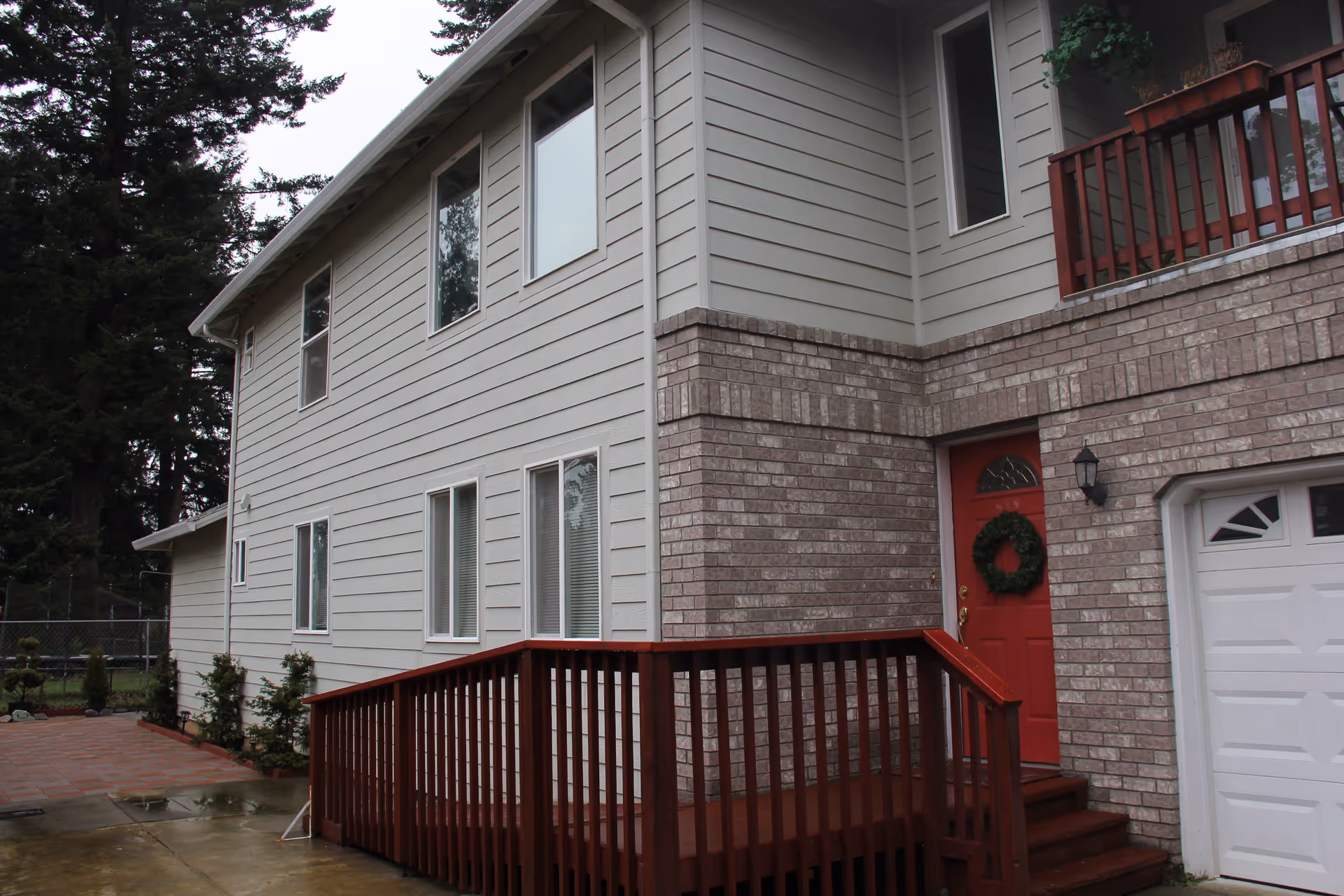 Exterior view of a two-story building with light gray siding and brick accents. There is a red door with a green wreath and a wooden ramp leading up to the entrance. A white garage door is visible on the right side, and tall trees are in the background.