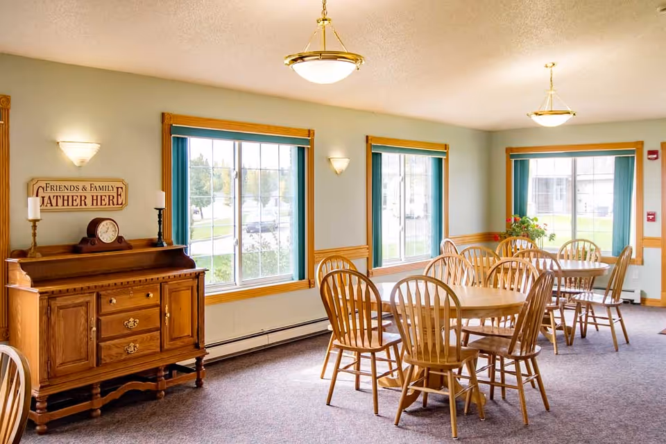 A bright dining room with multiple wooden tables and chairs arranged neatly. The room has large windows with green curtains allowing natural light to enter. A wooden sideboard with a clock and candles sits against one wall beneath a sign that reads 'Friends & Family Gather Here'. The walls are light-colored with wooden trim, and ceiling lights provide additional illumination.