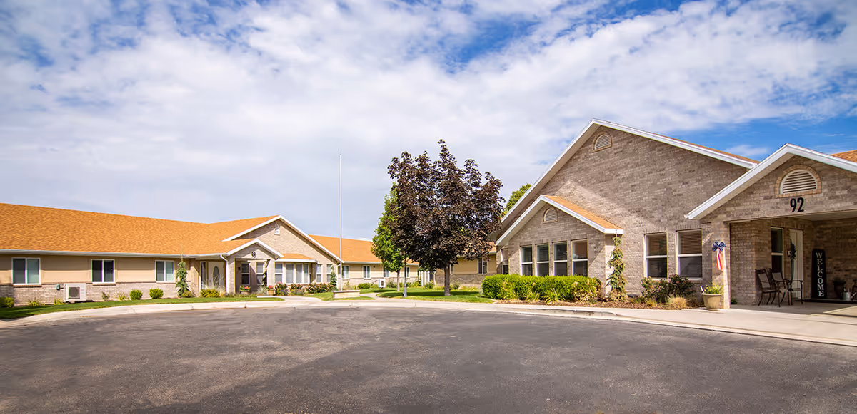 Exterior view of a single-story senior living facility with beige brick walls and orange shingle roofs under a partly cloudy sky. There is a circular driveway in front, a tree in the center, and a covered entrance with chairs and a welcome sign.