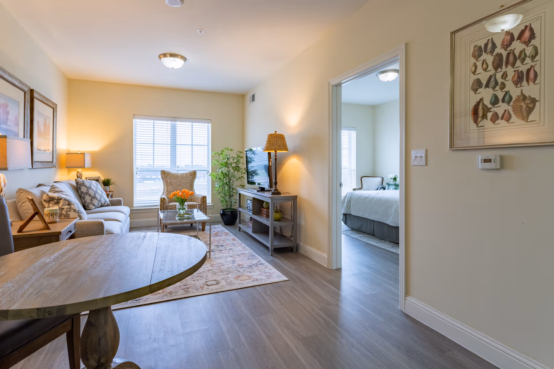 A bright and cozy living room with a beige sofa, patterned armchair, glass coffee table with a vase of orange flowers, a TV on a gray console, and a floor lamp. The room has light-colored walls, wood flooring, and a large window with blinds. A round wooden dining table is partially visible in the foreground. An open doorway leads to a bedroom with a bed, chair, and window.