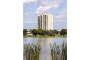 Tall residential building sits near a lake with trees and reeds in the foreground.
