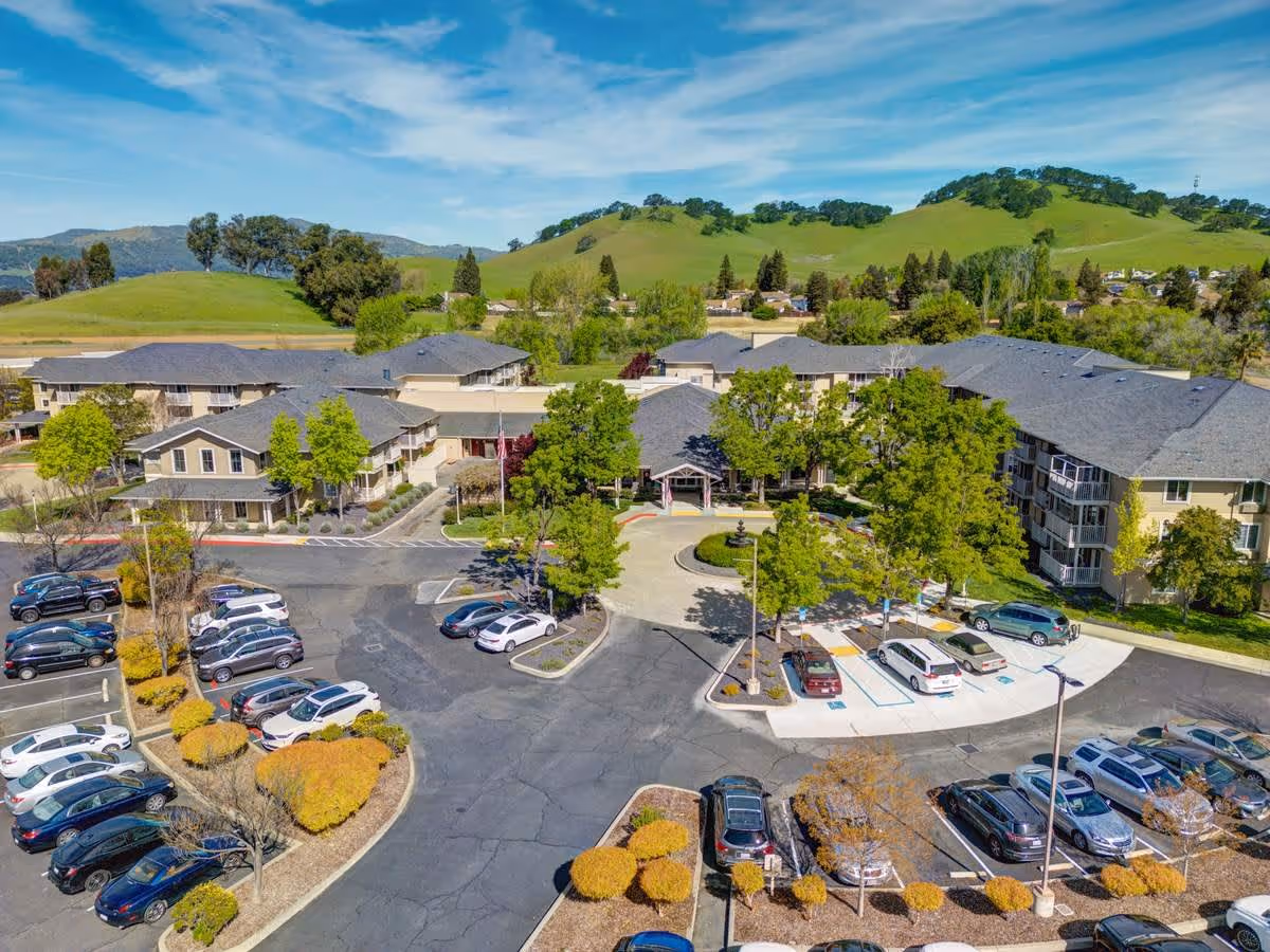 Aerial view of The Village at Rancho Solano senior living facility showing multiple buildings surrounded by trees and greenery, with a parking lot filled with cars in the foreground and rolling green hills in the background under a blue sky with scattered clouds.
