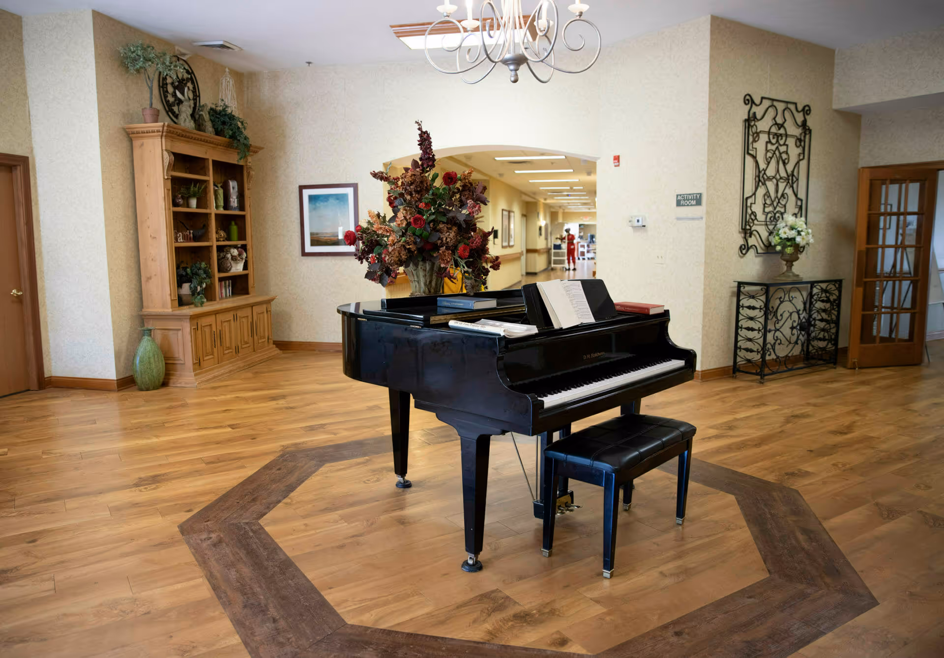 Interior view of a senior living facility featuring a black grand piano with an open music book on it, placed on a wooden floor with a decorative inlay. Behind the piano is a large floral arrangement. The room has light-colored walls, a wooden cabinet with plants and decorative items on the left, and a metal decorative wall piece with a vase of flowers on the right. A hallway labeled 'Activity Room' is visible in the background.
