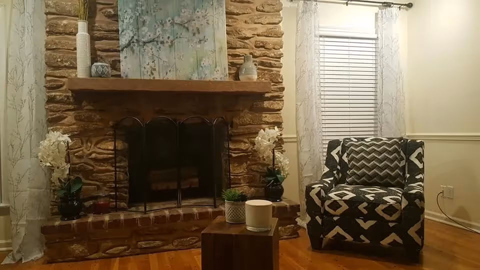 Cozy living room area with a stone fireplace in the center, decorated with vases and a painting on the mantel. To the right, there is a patterned armchair with a matching pillow, next to a window with white blinds and sheer curtains. A small wooden table in front of the fireplace holds a candle and a small plant.