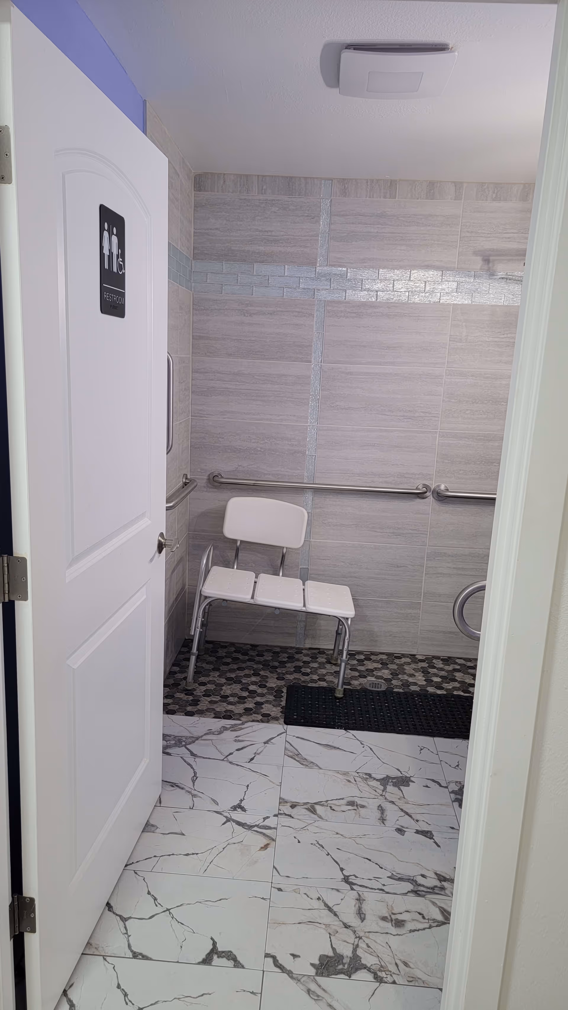 View into an accessible restroom with a white door partially open showing a sign for restroom with male, female, and wheelchair symbols. Inside, there is a white shower chair with a backrest against a tiled wall with gray and blue decorative tiles. The floor has white marble-patterned tiles transitioning to small hexagonal dark tiles in the shower area with a black mat.