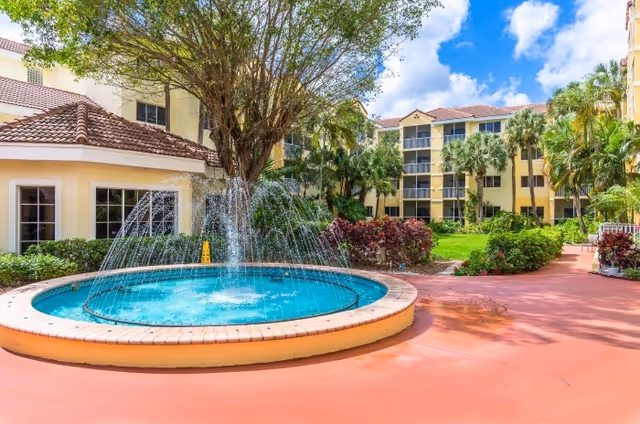 Sunny courtyard featuring a circular fountain, palm trees, and tan residential buildings.