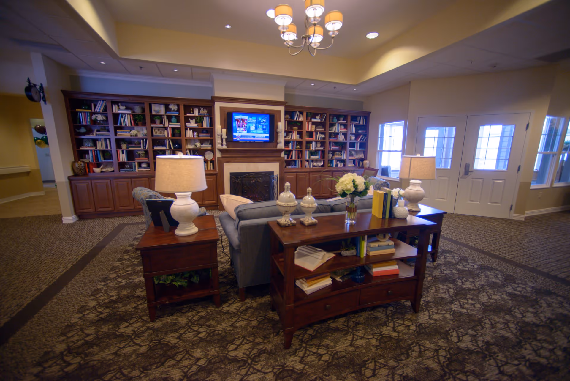 A cozy living room area in a senior living facility featuring a large wooden bookshelf filled with books and decorative items, a fireplace with a TV mounted above it, a gray sofa, wooden side tables with lamps, and a wooden console table with books and decorative vases. The room has carpeted flooring and soft lighting from ceiling fixtures.