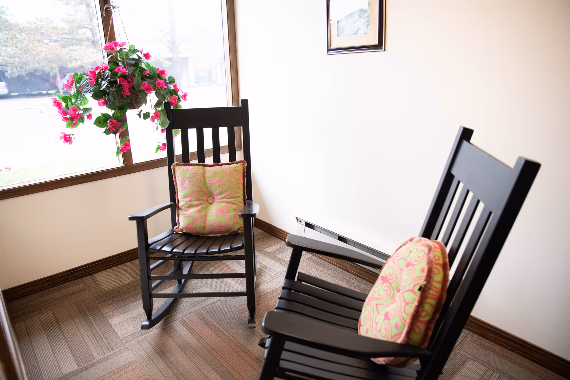 Two black wooden rocking chairs with patterned cushions are placed in a corner near a large window. A hanging basket with vibrant pink flowers is suspended near the window, and a framed picture is mounted on the white wall above a baseboard heater.