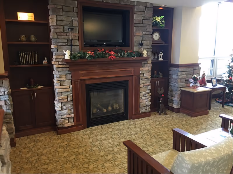 Cozy living room area with a stone and wood fireplace decorated with garland and small figurines. Built-in wooden shelves flank the fireplace, holding books, a clock, and decorative items. A TV is mounted above the fireplace. The room has carpeted flooring and a window letting in natural light. A cushioned wooden chair is partially visible in the foreground.
