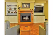 A cozy living room area featuring a wooden fireplace with a framed painting above it, flanked by built-in shelves and cabinets. Two gray upholstered chairs face the fireplace with a small wooden table between them. The floor is wooden, and a flat-screen TV is mounted in the right built-in cabinet.
