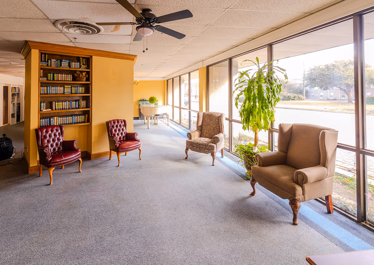 Bright senior living lounge with upholstered chairs, a tall bookshelf, and large windows along one wall.