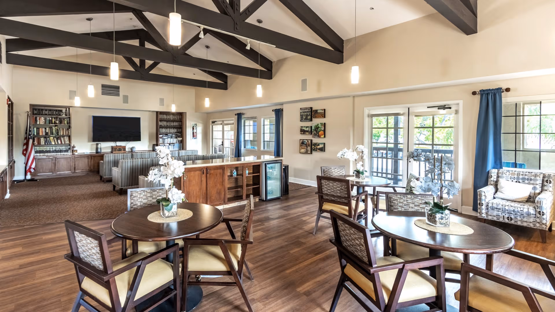 A spacious senior living common area with wooden floors, round tables with chairs, floral centerpieces, a seating area with striped sofas, a large flat-screen TV mounted on the wall, bookshelves, and large windows with blue curtains letting in natural light.