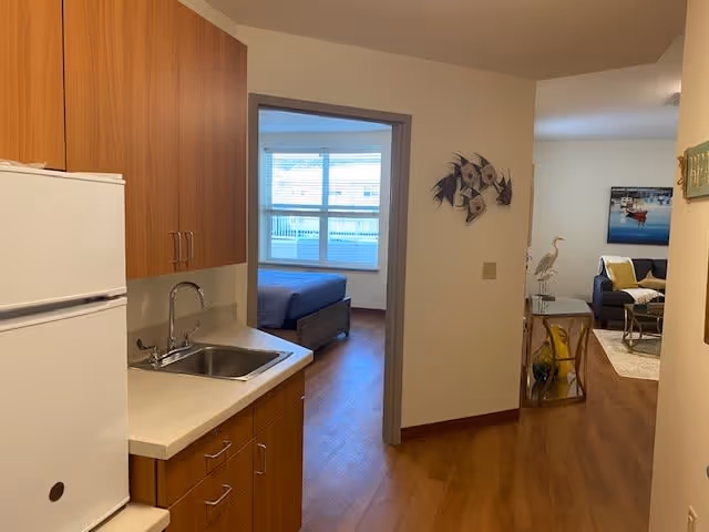 View of a small kitchen area with wooden cabinets, a white refrigerator, and a sink. Beyond the kitchen, there is a bedroom with a bed and large windows. To the right, there is a living room area with a sofa, a side table with a decorative bird sculpture, and wall art.