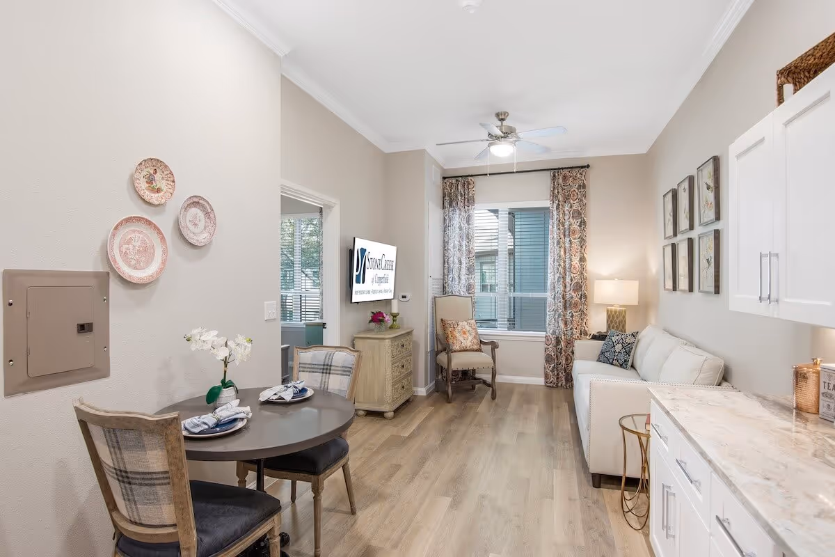A bright and cozy senior living room with light wood flooring, a small round dining table with two plaid chairs, a beige armchair near a window with patterned curtains, a white sofa with decorative pillows, white cabinets with a marble countertop, and a ceiling fan with a light. A TV mounted on the wall displays the StoneCreek of Copperfield Senior Living logo.