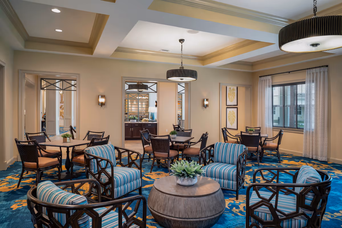 A well-lit common area in a senior living facility featuring a seating arrangement with four striped armchairs around a round wooden coffee table with a small plant. In the background, there are multiple tables with chairs, decorative wall art, and large windows with sheer curtains. The room has a blue and yellow patterned carpet and modern ceiling lights.