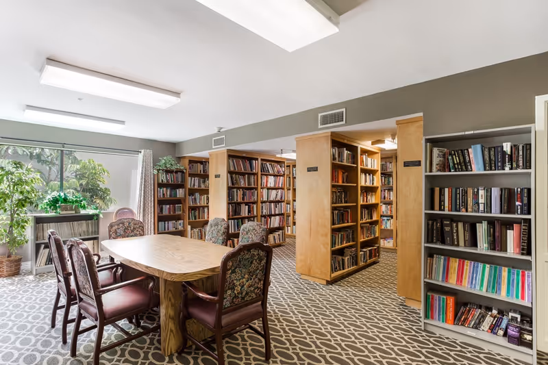 A well-lit library room with wooden bookshelves filled with books, a wooden table surrounded by six upholstered chairs with floral patterns, a large window with curtains, and a potted plant near the window. The floor is carpeted with a geometric pattern.