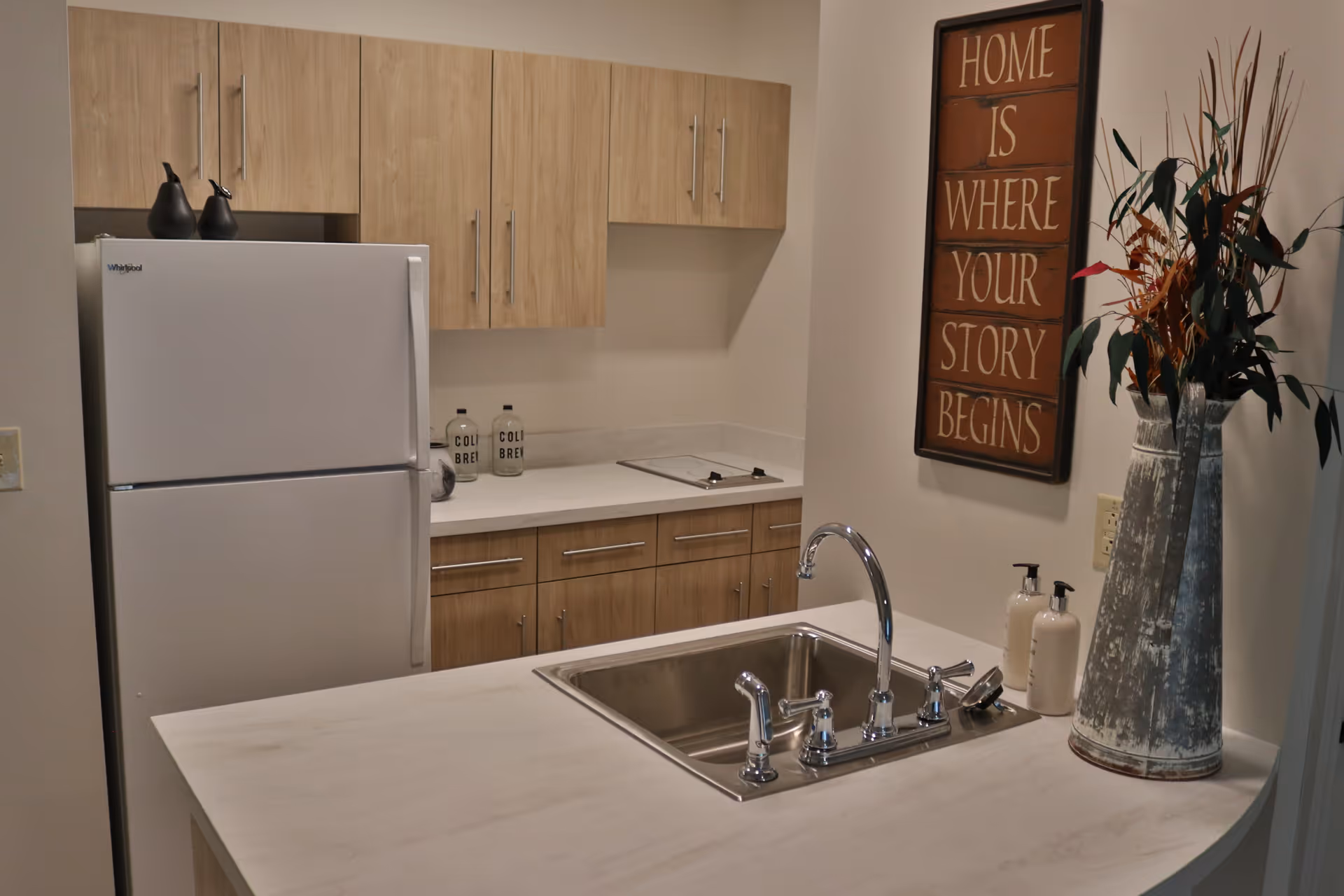 A modern kitchen area with light wood cabinets, a white Whirlpool refrigerator, a stainless steel sink with a chrome faucet on a white countertop, two soap dispensers, and a decorative vase with tall plants. On the wall, there is a framed sign that reads 'HOME IS WHERE YOUR STORY BEGINS.'