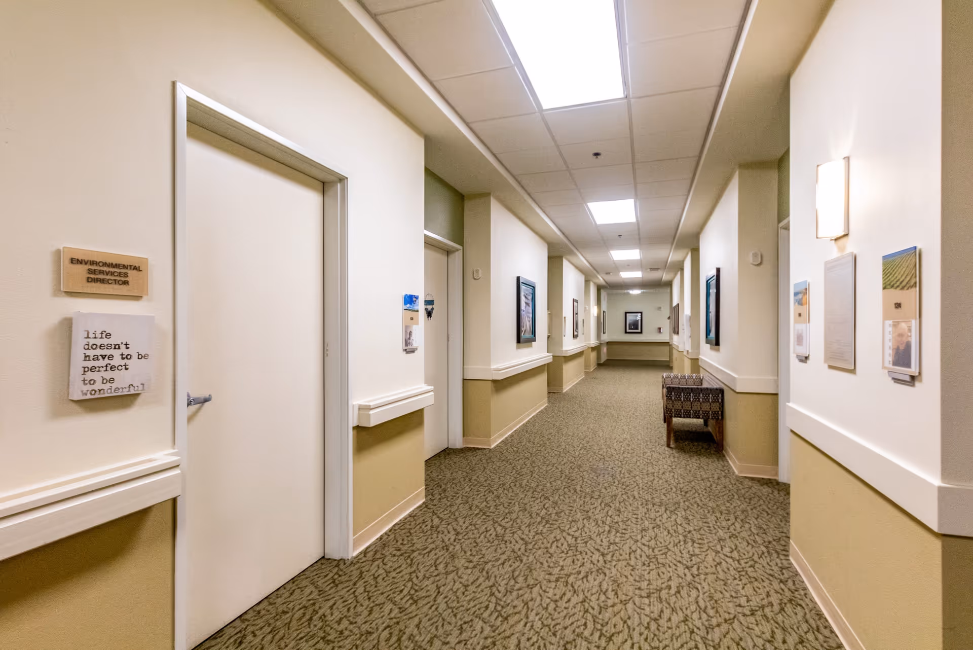 A long, well-lit hallway in a senior living facility with beige walls and patterned carpet. Several doors and framed pictures line the walls, and a single chair is placed along the right side. A sign on the left door reads 'Environmental Services Director' and a decorative plaque below it says 'life doesn't have to be perfect to be wonderful.'