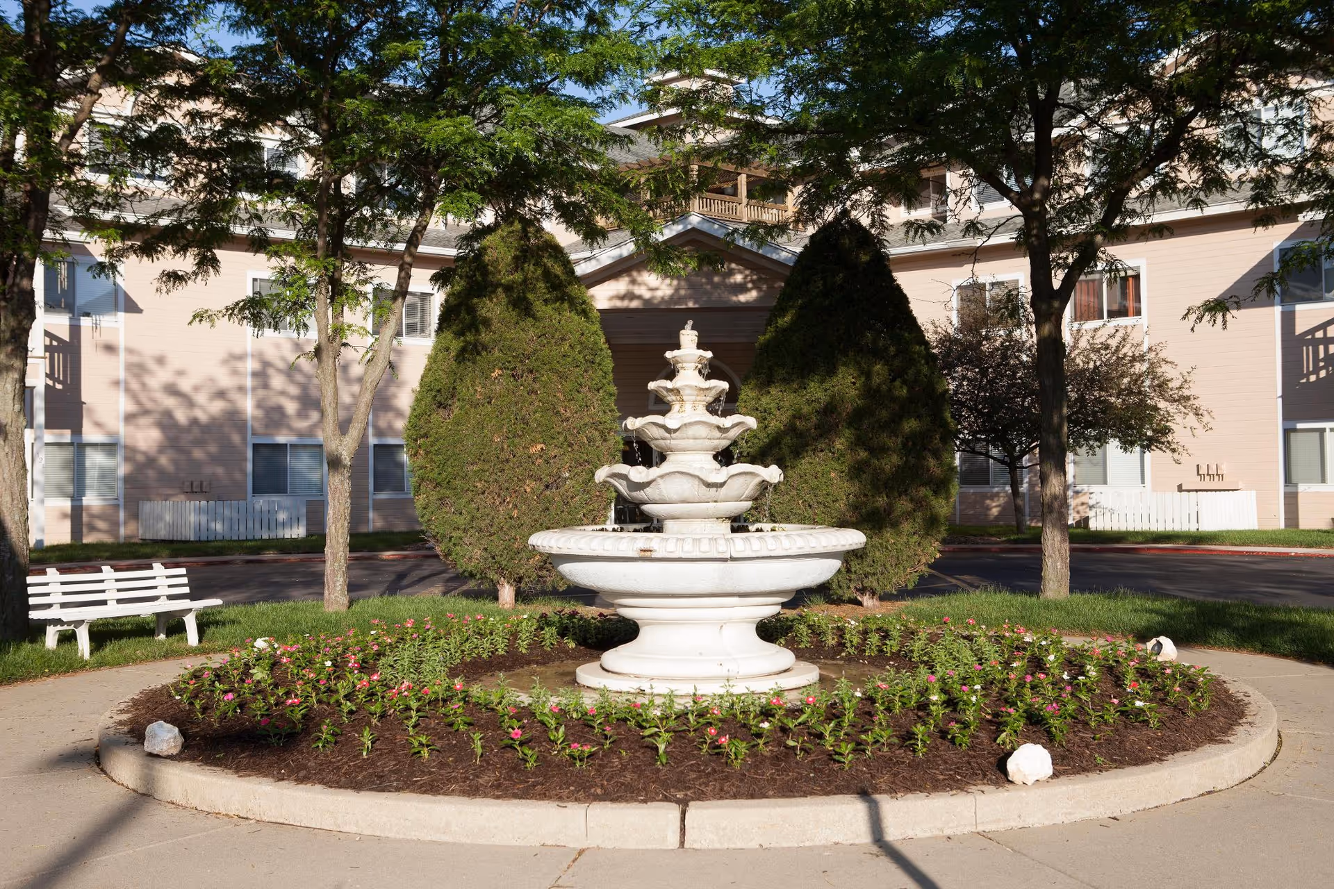 A white tiered water fountain surrounded by a circular flower bed with small pink flowers and mulch, located in front of a multi-story beige building. There are two large conical evergreen bushes behind the fountain, along with several trees and a white bench to the left side of the image.