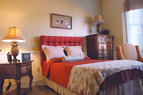 A cozy bedroom featuring a red-upholstered bed with pillows and a throw, nightstands with lamps, a dresser, and an armchair by a window.