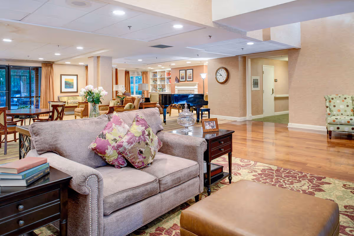 A spacious and well-lit living room area in a senior living facility featuring a beige loveseat with floral pillows, a wooden side table with books and a photo frame, a large patterned rug, a brown ottoman, and a grand piano in the background. The room has wooden flooring, beige walls, multiple seating areas, framed artwork, and large windows with curtains allowing natural light.