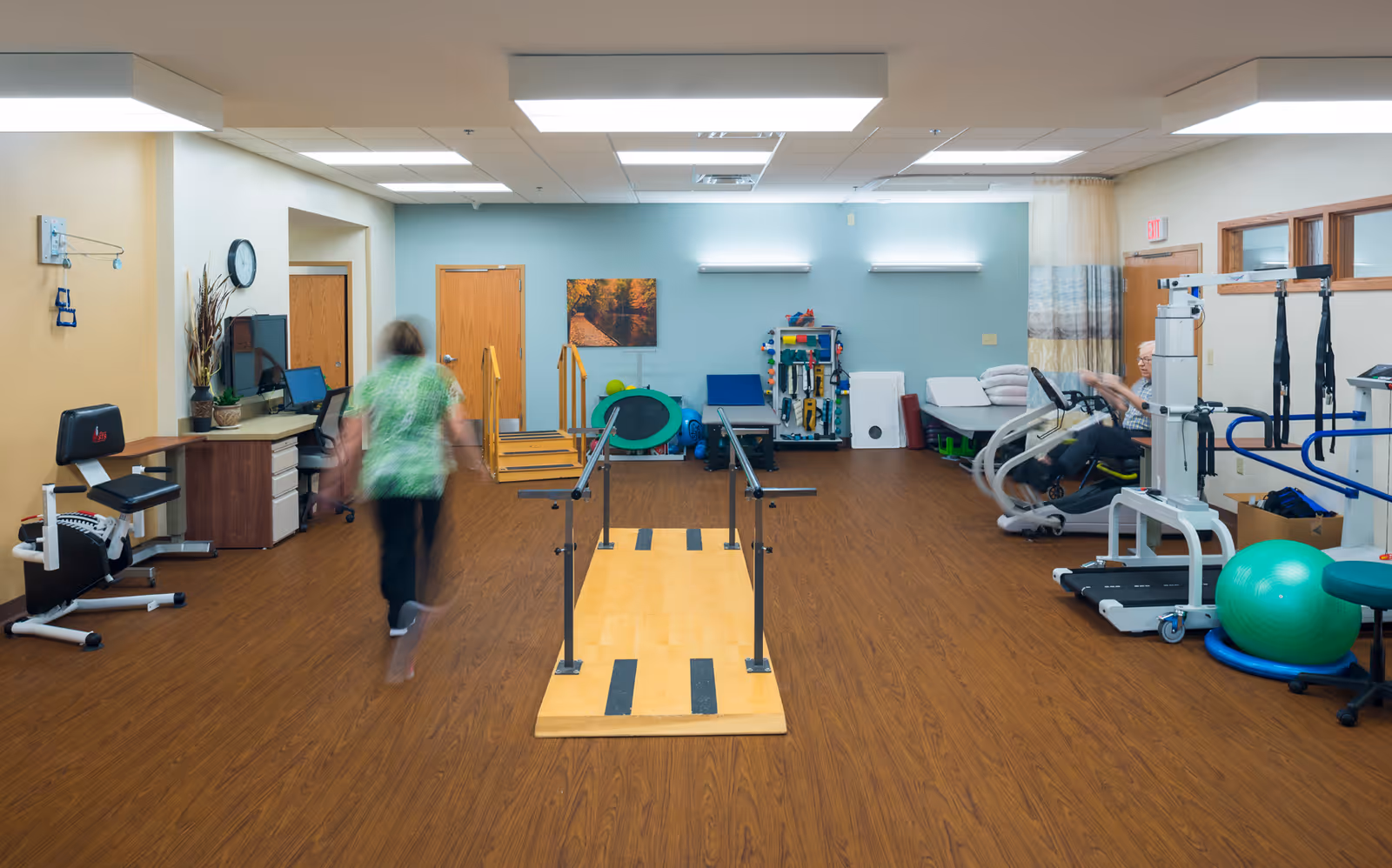 A rehabilitation or physical therapy room with wooden flooring and light blue walls. The room contains various exercise and therapy equipment including parallel bars, exercise balls, a stationary bike, and other physical therapy tools. A person in a green shirt is walking towards the back of the room, and another person is seated on a piece of exercise equipment on the right side. There is a desk with a computer on the left side and a door at the back wall.