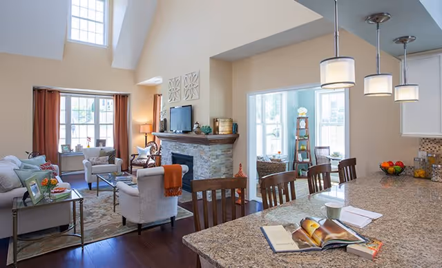 Bright and spacious living room area with high ceilings, featuring a stone fireplace with a TV mounted above it. The room has beige walls, large windows with brown curtains, and comfortable seating including armchairs and a sofa. Adjacent to the living room is a kitchen area with a granite countertop island, wooden chairs, and pendant lights hanging above. The kitchen counter has an open book, a cup, and a bowl of fruit. A sunroom with large windows and additional seating is visible in the background.
