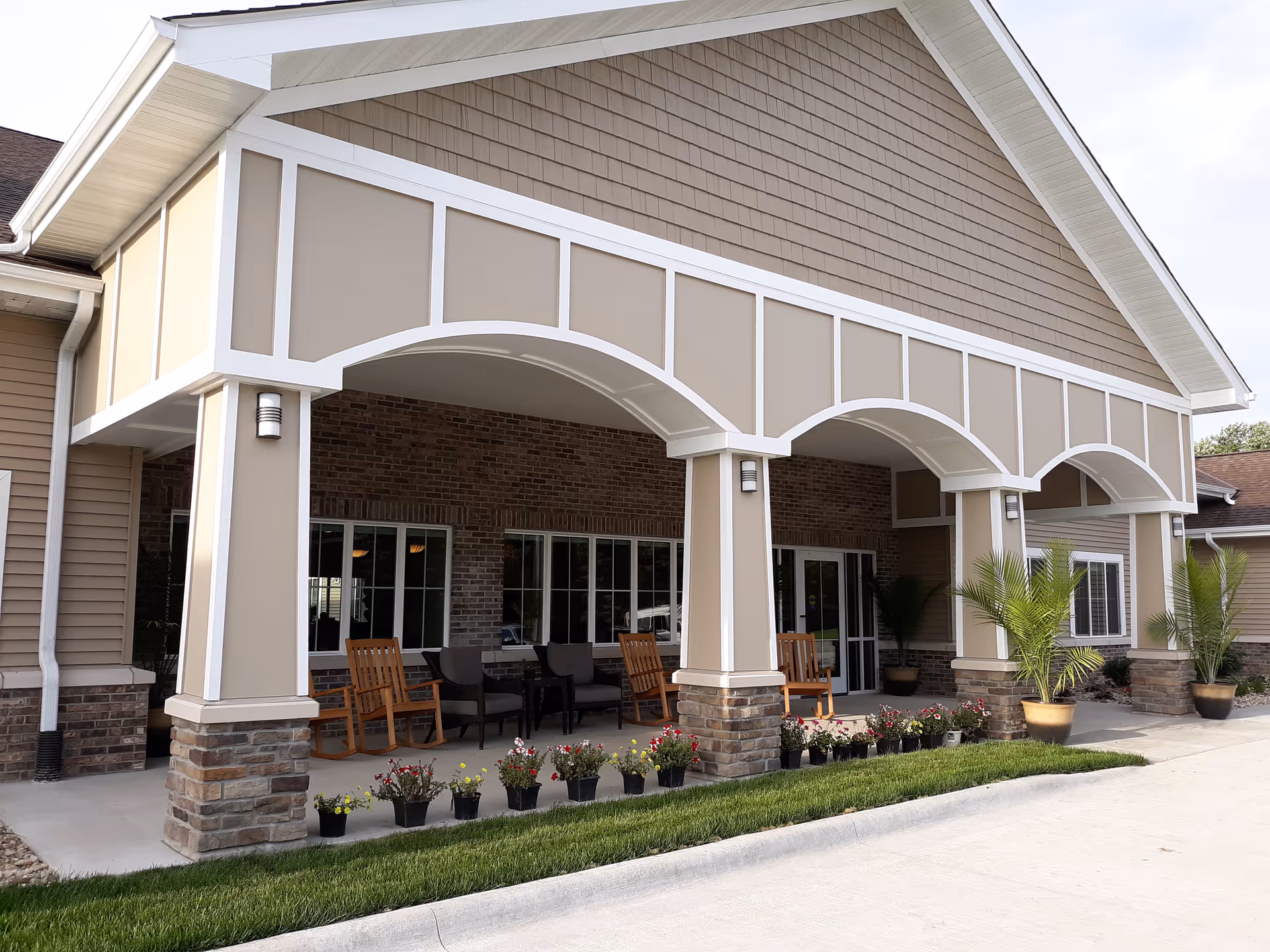 Exterior view of the entrance to a senior living facility with a covered porch area featuring beige and white trim, stone pillars, several potted plants, rocking chairs, and cushioned chairs arranged for seating.