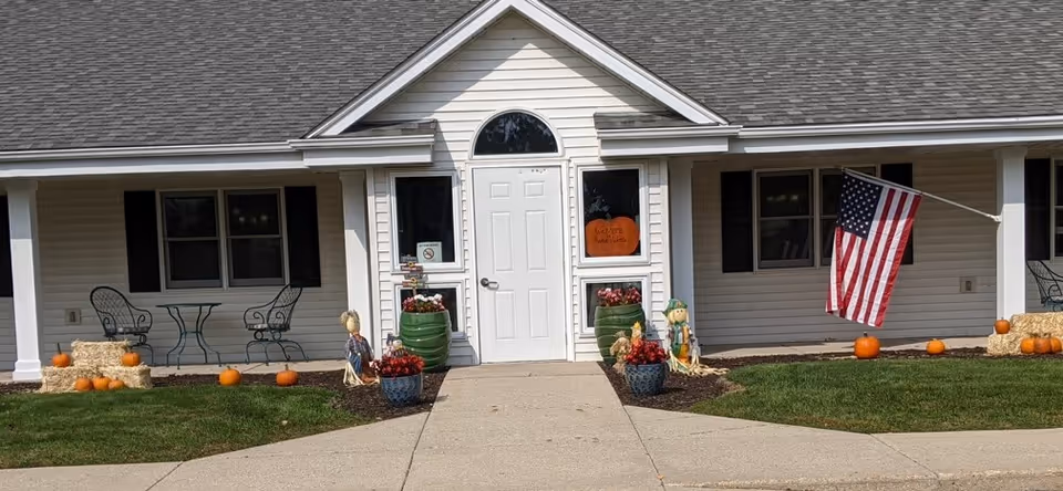 Front exterior view of a single-story building with white siding and a gray shingled roof. The entrance features a white door with two windows on either side, decorated with fall-themed items including pumpkins, hay bales, and scarecrows. An American flag is mounted on the right side of the building. There is a small patio area with a metal table and chairs on the left side.