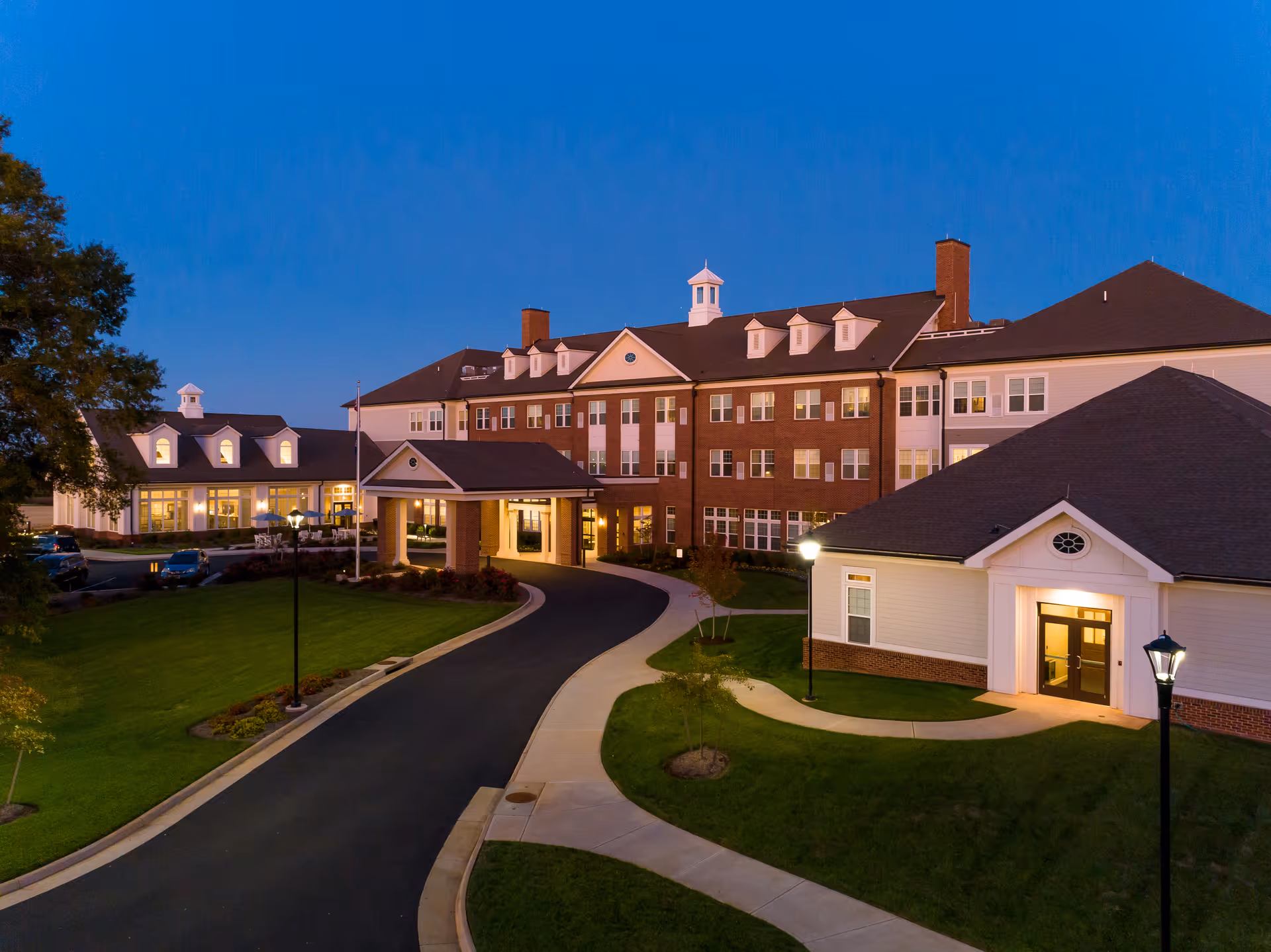 Exterior view of The Culpeper senior living facility at dusk, showing a large, multi-story brick and white building with a covered entrance, well-lit pathways, and landscaped green lawns.