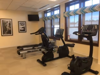 A small fitness room with exercise equipment including two stationary bikes and two treadmills. The room has beige walls, three windows with patterned valances, a wall-mounted TV, and two framed black and white pictures on the wall.