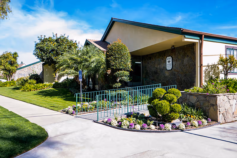 Exterior view of Catered Manor Nursing Center showing a single-story building with stone and stucco walls, a ramp with green railings, landscaped garden beds with shrubs and flowers, and a clear blue sky.