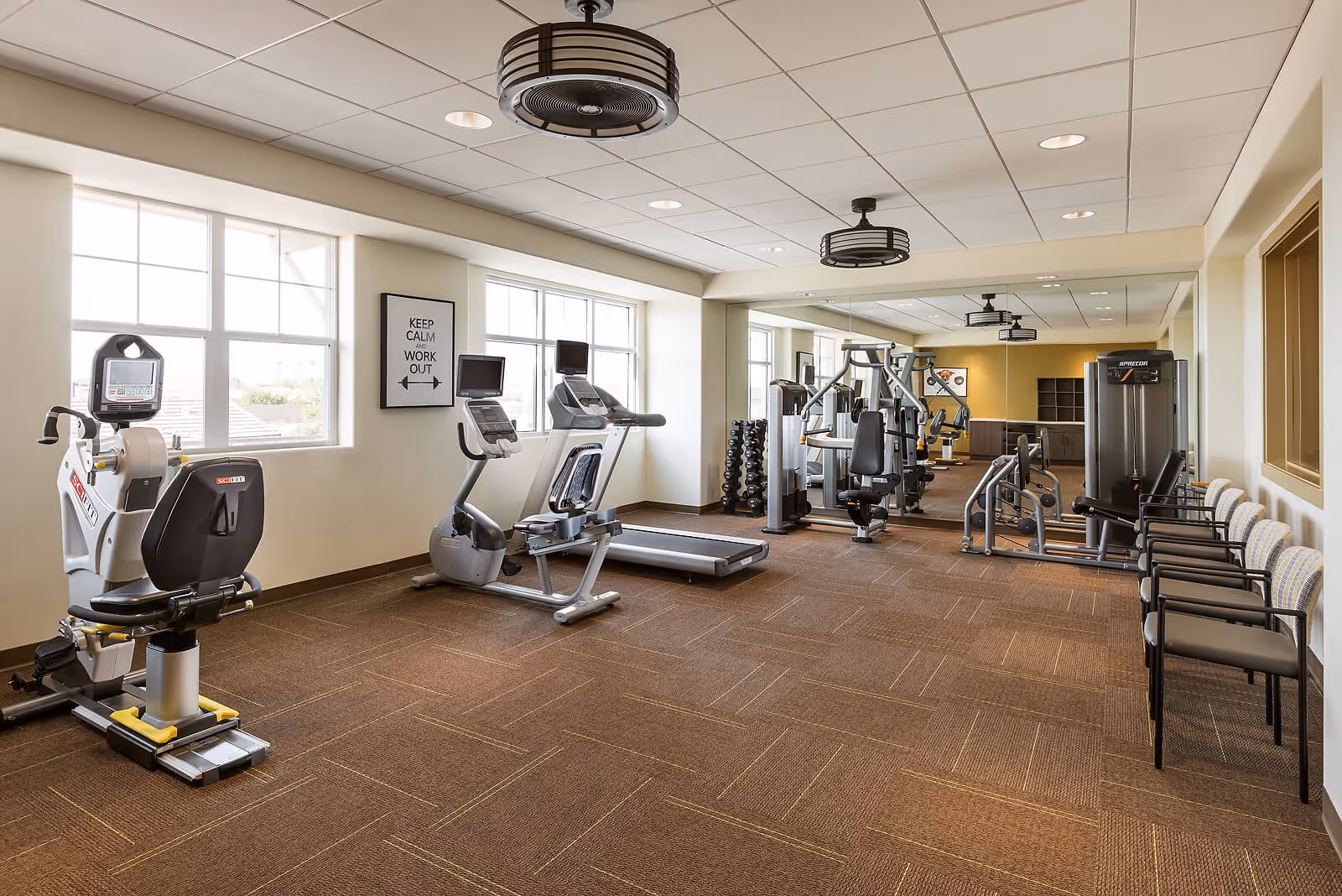 A well-lit fitness room with exercise equipment including a recumbent bike, treadmill, weight machines, and a rack of dumbbells. There are large windows letting in natural light, a motivational sign on the wall that reads 'KEEP CALM AND WORK OUT', and several chairs lined up against the wall. The room has a brown carpeted floor and a ceiling with recessed lighting and circular ceiling fans.