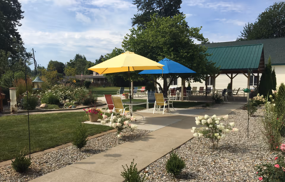 Outdoor patio area at Union Court Assisted Living with a concrete walkway, colorful patio chairs, and tables shaded by yellow and blue umbrellas. The area is surrounded by landscaped garden beds with flowers and shrubs, and a covered pavilion with additional seating is visible in the background under a green roof.