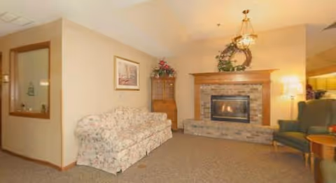 A cozy living room with a floral patterned sofa, a green armchair, a brick fireplace with a wooden mantle decorated with a wreath and flowers, a floor lamp, and a framed picture on the wall.