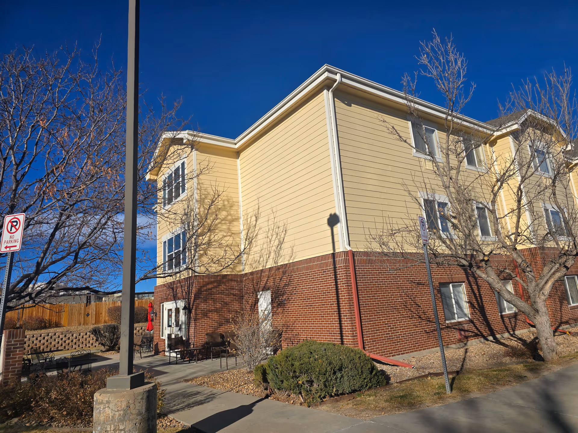 Three-story yellow-and-brick assisted living building exterior with leafless trees, benches, and a sidewalk under a clear blue sky.