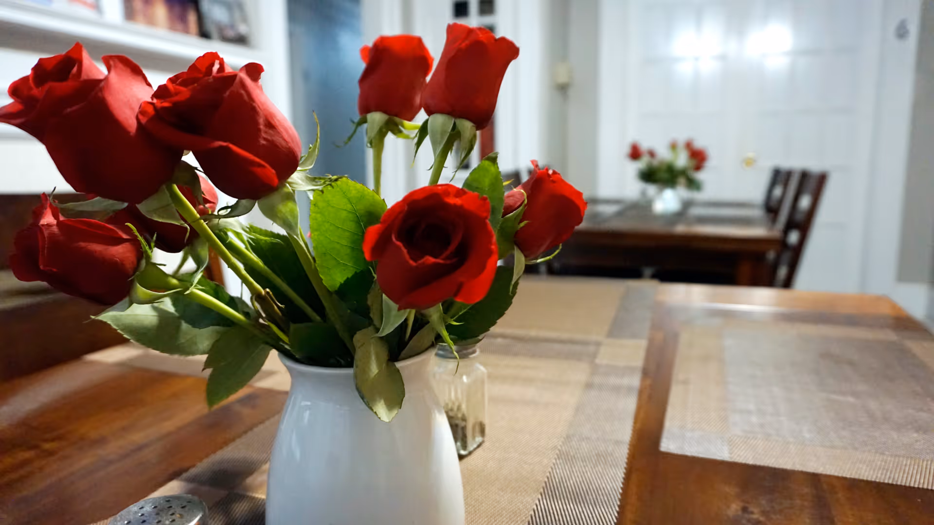 Close-up of a white vase with red roses on a wooden dining table with placemats. In the background, another dining table with a similar vase and red roses is visible in a softly lit room.