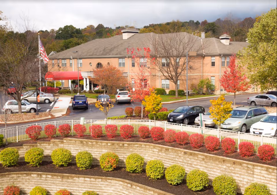 Front view of a two-story brick senior living building with landscaped terraced beds, parked cars, and an American flag.