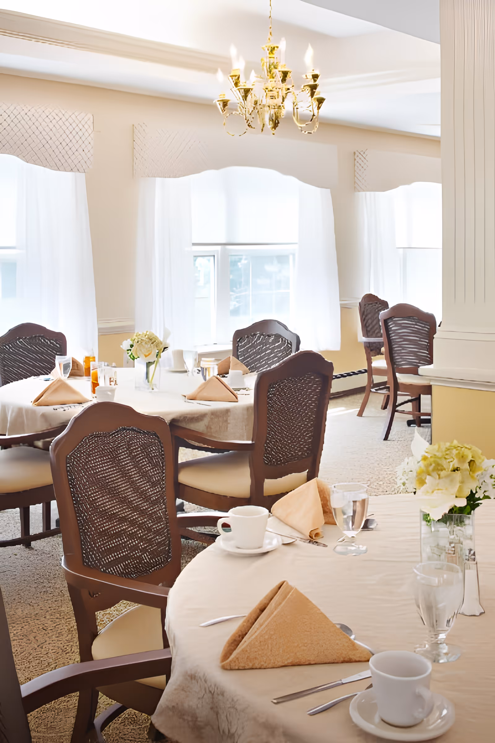 A bright dining room with round tables covered in beige tablecloths, each set with folded napkins, cups, glasses, and silverware. The room features wooden chairs with woven backs, large windows with white curtains, a decorative column, and a gold chandelier hanging from the ceiling.