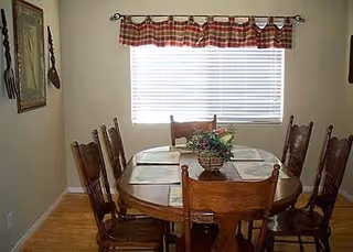 A dining room with a wooden oval table surrounded by six wooden chairs. The table has placemats and a centerpiece with a plant. A window with white blinds and a red and white checkered valance is on the wall behind the table. The walls are light-colored, and there is a framed picture and a hanging decoration on the left wall.