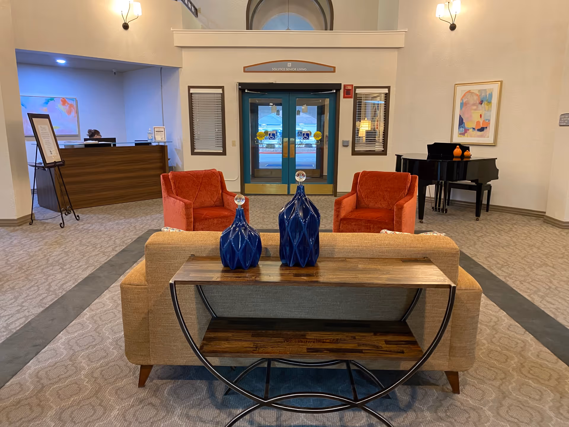 Interior view of a senior living facility lobby with a beige sofa and two red armchairs arranged around a wooden console table with two blue decorative vases. In the background, there is a reception desk with a person sitting behind it, double glass doors with handicap accessibility signs, a black grand piano, and colorful abstract artwork on the walls.