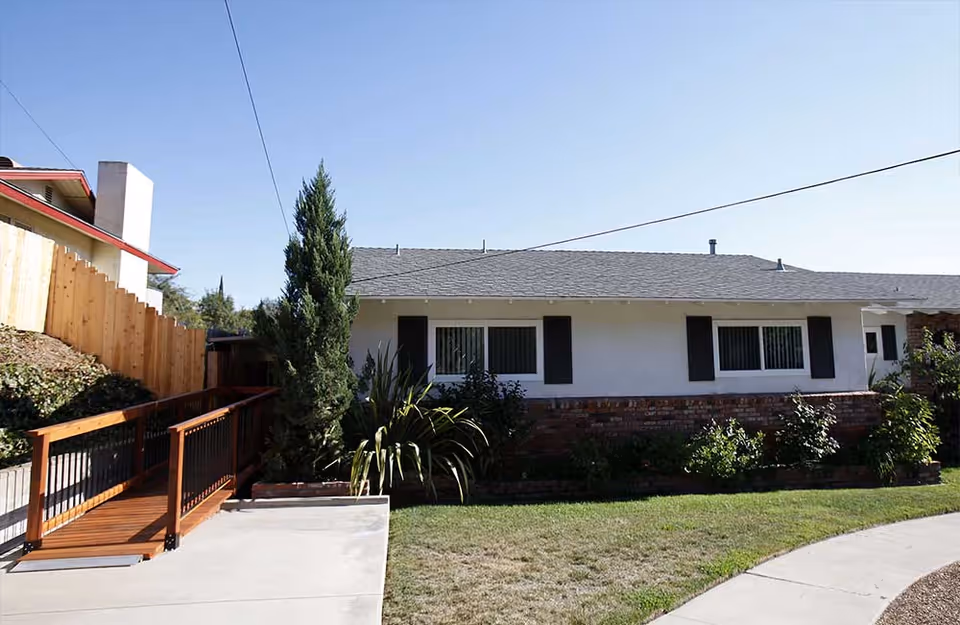 Exterior view of a single-story residential building with white walls, black window shutters, and a gray shingled roof. There is a wooden ramp with black railings leading up to the entrance on the left side. The building is surrounded by a small garden with bushes and a tree, and there is a concrete pathway and lawn in the foreground under a clear blue sky.