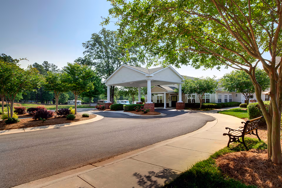 Driveway entrance with a covered porte-cochère, landscaped grounds, and a bench outside a senior living facility.