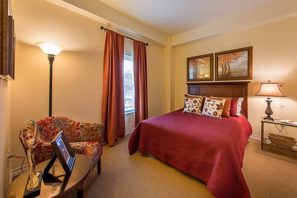 Cozy furnished bedroom with a red bedspread, patterned armchair, window with curtains, lamps, and framed artwork above the headboard.