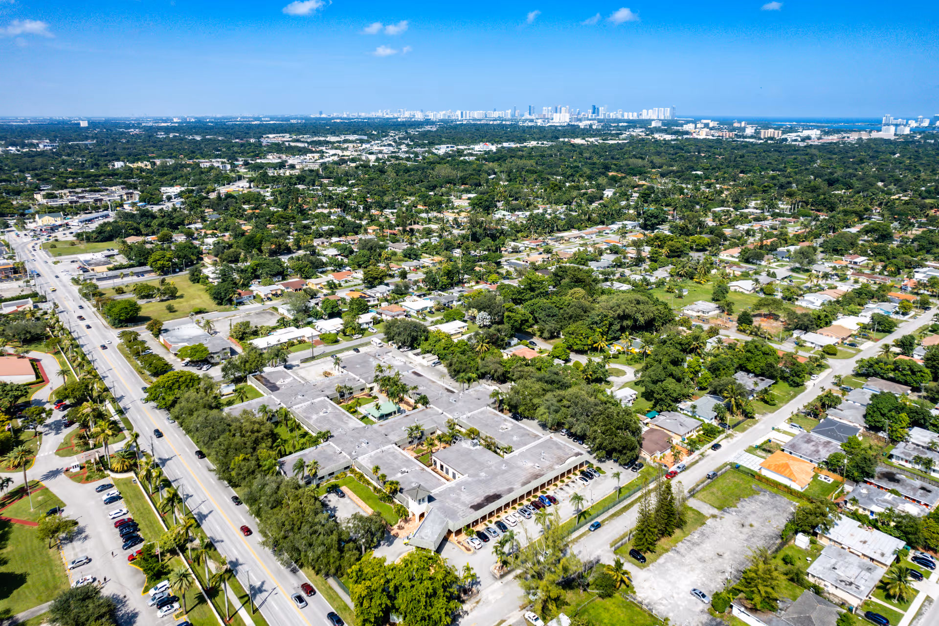 Aerial view of a suburban area with a large building complex surrounded by trees and residential houses. The image shows a wide road with cars parked along it and a distant city skyline under a clear blue sky.